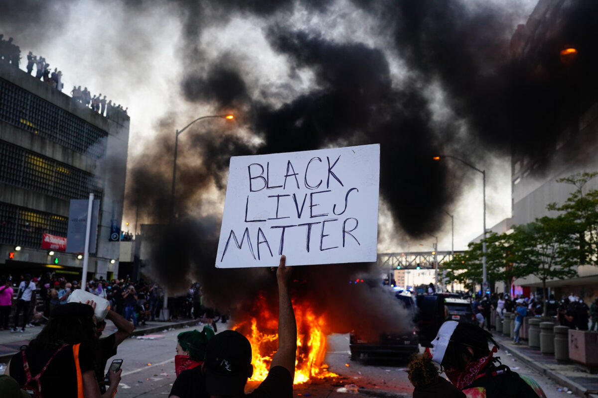 Một người đàn ông cầm tấm biển BLM khi một chiếc xe cảnh sát bốc cháy trước mặt anh trong cuộc biểu tình bên ngoài Trung tâm CNN ở Atlanta, Georgia, vào ngày 29/5/2020. (Ảnh Elijah Nouvelage/ Getty Images)