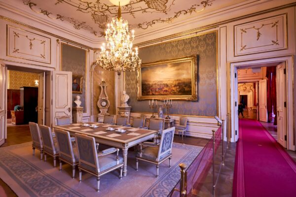 Phòng hội nghị, cũng tọa lạc bên trong Các dãy phòng Hoàng gia, là một cách tiếp cận thận trọng hơn với phong cách kiến trúc Baroque. Một chiếc đèn chùm pha lê bằng vàng treo trên trần nhà, hoa văn bằng thạch cao mịn, cùng những bức tường và khung viền cửa với họa tiết trang trí mạ vàng. Tại đây màu xanh lam là tông màu chủ đạo, hiện hữu xuyên suốt trên tấm thảm trải sàn, thảm treo tường, và những chiếc ghế có tông màu-xanh-lam-và-xám-bạc trang nhã. (Ảnh: Marcobrivio.photo/Shutterstock)