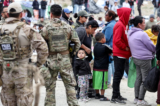 Các nhân viên và quan chức chấp pháp liên bang đang theo dõi trong khi những người nhập cư xếp hàng để được chở đi từ một trại tạm bợ giữa các bức tường biên giới Hoa Kỳ-Mexico ở San Diego hôm 13/05/2023. (Ảnh: Mario Tama/Getty Images)