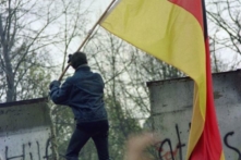 Một người dân Tây Đức mang theo lá cờ của Cộng hòa Liên bang Đức trao cho người dân Đông Đức qua một phần Bức tường Berlin sụp đổ, gần Cánh Cổng Brandenburg vào ngày 11/11/1989. Ba nhà lãnh đạo là cựu Tổng thống Hoa Kỳ Ronald Reagan, nữ Thủ tướng Anh Margaret Thatcher, và Đức Giáo Hoàng Ba Lan John Paul II, đã góp phần quan trọng vào sự sụp đổ của Bức tường Berlin, đồng thời kết thúc Chiến Tranh Lạnh. (Ảnh: Gerard Malie/AFP via Getty Images)