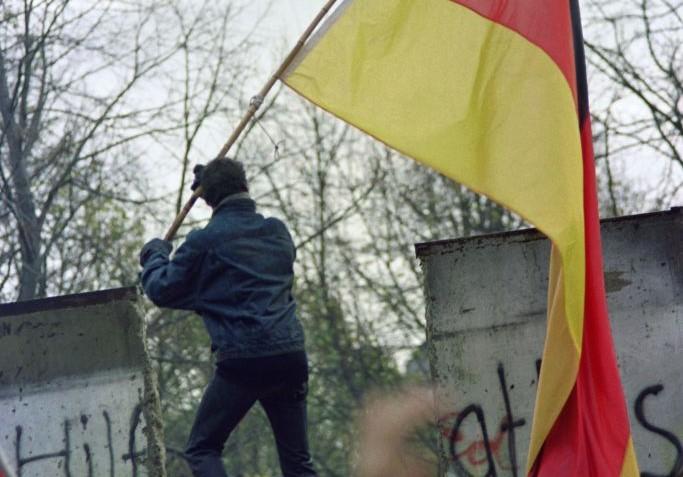 Một người dân Tây Đức mang theo lá cờ của Cộng hòa Liên bang Đức trao cho người dân Đông Đức qua một phần Bức tường Berlin sụp đổ, gần Cánh Cổng Brandenburg vào ngày 11/11/1989. Ba nhà lãnh đạo là cựu Tổng thống Hoa Kỳ Ronald Reagan, nữ Thủ tướng Anh Margaret Thatcher, và Đức Giáo Hoàng Ba Lan John Paul II, đã góp phần quan trọng vào sự sụp đổ của Bức tường Berlin, đồng thời kết thúc Chiến Tranh Lạnh. (Ảnh: Gerard Malie/AFP via Getty Images)