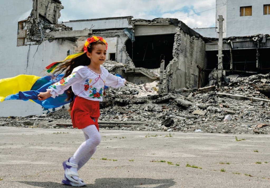 Một bé gái cầm quốc kỳ Ukraine chạy trước một trung tâm văn hóa bị hư hại trong lễ tốt nghiệp của sinh viên nghệ thuật ở thị trấn Derhachi, vùng Kharkiv, miền đông Ukraine. (Ảnh: Sergey Bobok/Cộng tác viên/AFP/Getty Images)