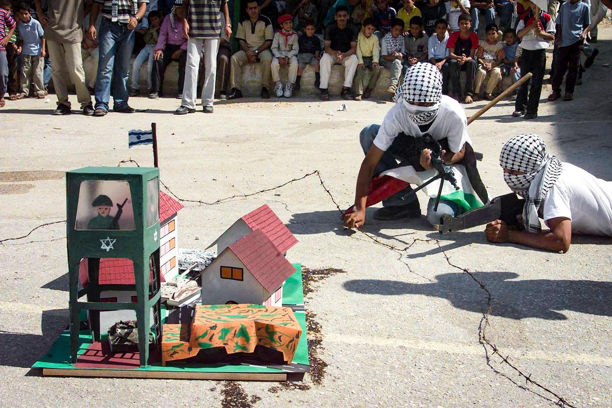 Trẻ em và các bậc cha mẹ theo dõi những cậu bé người Palestine đeo khăn trùm đầu và cầm mô hình súng trường tự động khi các em thực hiện một cuộc tấn công giả vào một khu định cư của người Do Thái vào ngày cuối cùng của trại hè ở Tây Ngạn của thị trấn Nablus vào ngày 15/08/2002. (Ảnh: Jaafar Ashtiyeh/ AFP qua Getty Images)