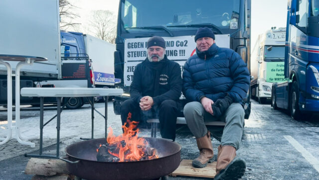 Hai nhân viên giao nhận hàng hóa và tài xế xe tải sưởi ấm bên bếp lửa trong cuộc biểu tình. (Ảnh: Matthias Kehrein/Epoch Times)